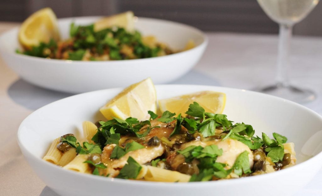 two bowls of chicken piccata with a glass of wine in the background